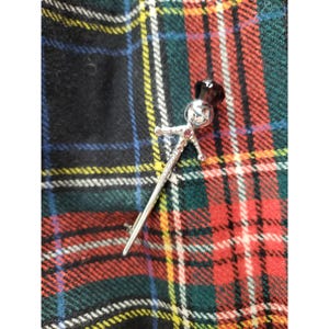 Op de afbeelding: Zilveren kiltspeld met een decoratief figuur aan de bovenkant, met een gezicht en een donkere edelsteen op de kop. De speld heeft kleine rode edelstenen op het lichaam en de armen. De achtergrond is een tartanpatroon in rood, groen, zwart en geel.