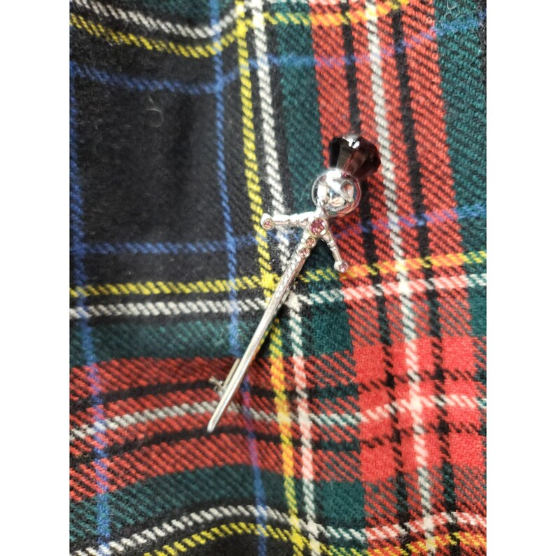 Op de afbeelding: Zilveren kiltspeld met een decoratief figuur aan de bovenkant, met een gezicht en een donkere edelsteen op de kop. De speld heeft kleine rode edelstenen op het lichaam en de armen. De achtergrond is een tartanpatroon in rood, groen, zwart en geel.