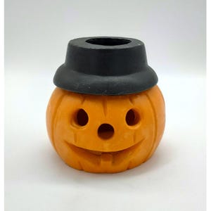 May include: A bright orange jack-o'-lantern candle holder shaped like a round pumpkin with a carved face featuring two circular eyes, a small circular nose, and a wide smiling mouth, topped with a solid black hat-shaped lid.