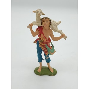 May include: A figurine of a shepherd carrying a lamb on his shoulders. The shepherd wears blue pants, a red and yellow patterned shawl, and a green bag. The lamb is white, and the figurine stands on a green base.