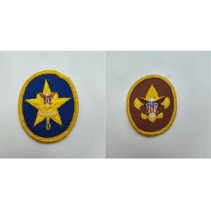May include: Two embroidered circular patches with yellow stitched borders. The left patch features a yellow star on a blue background with an eagle emblem. The right patch displays a yellow fleur-de-lis symbol on a brown background with an eagle.