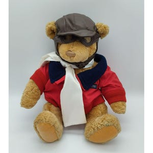 May include: A plush teddy bear wearing a brown aviator cap with goggles, a red jacket with a navy blue collar, and a white scarf. The bear is sitting with its arms at its sides. The jacket has the word "EXT" on the sleeve.