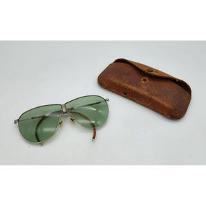 May include: Vintage aviator sunglasses with green lenses and a silver frame, next to a brown leather case. The sunglasses have a classic design, with a double bridge and teardrop-shaped lenses. The case is rectangular and has a button closure.