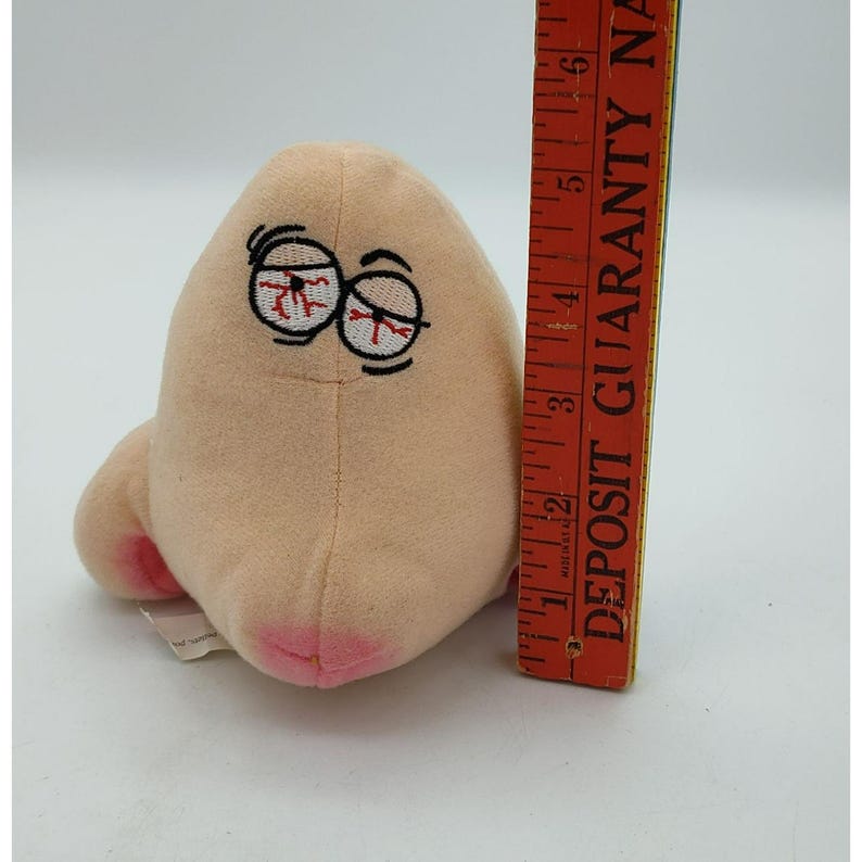 May include: A beige plush toy shaped like a nose with embroidered bloodshot eyes and pink nostrils. An orange ruler is positioned vertically next to the plush, showing a height of approximately 6 inches from the base to the top of the nose.