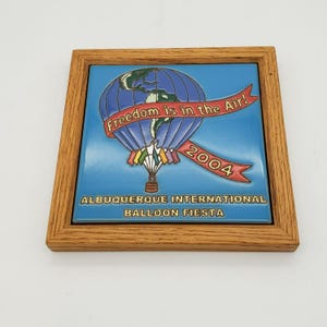 May include: A framed ceramic tile featuring a blue hot air balloon with the Earth design, the text "Freedom is in the Air!" and "2004". The tile is framed in a light brown wooden frame. The text "ALBUQUERQUE INTERNATIONAL BALLOON FIESTA" is at the bottom.