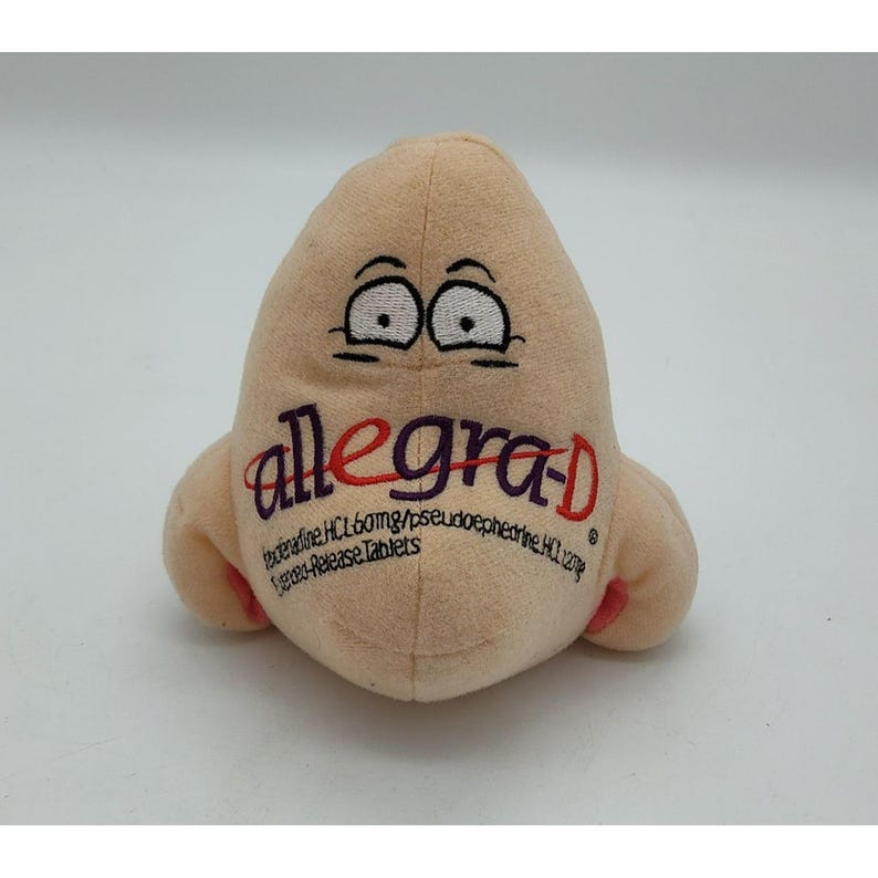 May include: A plush toy shaped like a human nose in a light beige color. The nose features embroidered black eyes with thick eyebrows and the text "allegra-D" in purple and red, along with smaller black text detailing medication ingredients.