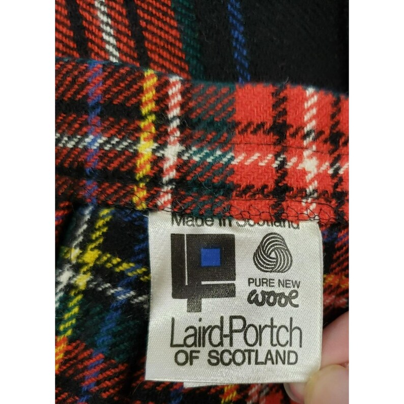 Op de afbeelding: Close-up van een rood, zwart, groen en wit geruite stof. Een label met de tekst "Made in Scotland" en het logo van Laird-Portch of Scotland, met de tekst "Pure New Wool".