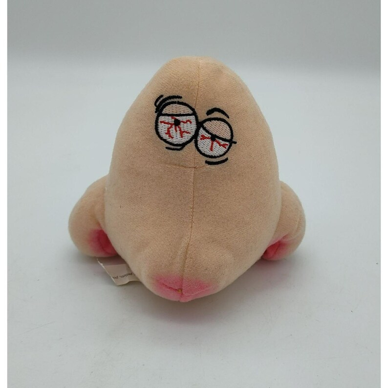May include: A plush toy shaped like a human nose with a light beige fabric exterior. The nose features embroidered eyes with red bloodshot details and pink-tinted nostrils at the base, creating a soft, stuffed anatomical representation.