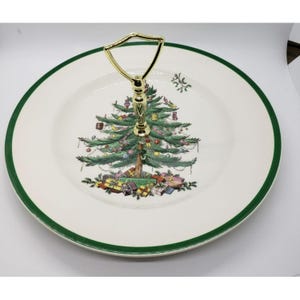 May include: A two-tiered serving plate with a gold handle. The plate features a Christmas tree design with ornaments and wrapped gifts. The rim has a green border. A festive piece for serving treats.