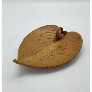May include: A tan ceramic dish shaped like a leaf with a curled stem at the top. The surface features a textured finish with darker brown speckles and embossed veins running from the center stem toward the edges of the leaf-shaped bowl.