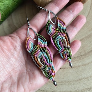 Puede incluir: Un par de pendientes de macramé hechos a mano en tonos verdes, azules, rosas, marrones y beige. Los pendientes tienen ganchos plateados y forma de lágrima. El intrincado anudado crea un diseño texturizado y estampado, ideal para un look bohemio.