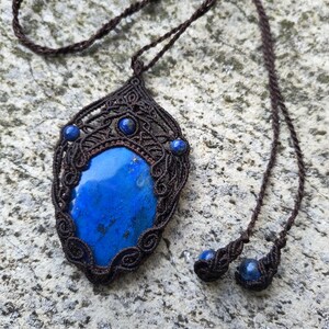 Puede incluir: Un collar con una gran piedra de lapislázuli azul en forma de lágrima, engastada en un intrincado trabajo de cordón de macramé marrón oscuro. El colgante está decorado con tres cuentas de lapislázuli más pequeñas y el cordón termina en dos cuentas a juego.