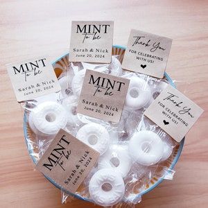 May include: A bowl of white mints with brown paper favor bags tied with twine. The bags have a white label with black text that reads "Mint to be" with the couple's names and wedding date. Some bags have a thank you label with a heart.