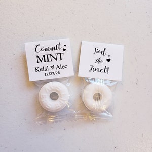 May include: Two white mint candies wrapped in clear plastic bags. Each bag has a small white card attached with black text. The card on the left says "Commit Mint" with the text "Kelsi ♥ Alec 12/27/26" below. The card on the right says "Tied the Knot!"