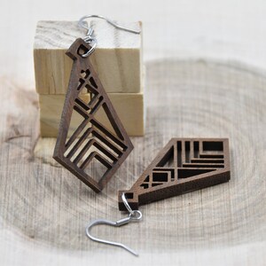 May include: Geometric wooden earrings with a dark brown finish. The earrings feature a unique open design with angular shapes and silver-colored ear wires. One earring is displayed separately, and the other is attached to a silver hook.
