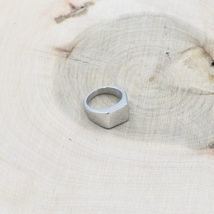 Peut inclure: Une bague argentée avec une face carrée. La bague est en métal brillant et repose sur une surface en bois clair. Le design de la bague est simple et moderne, adapté au port quotidien.
