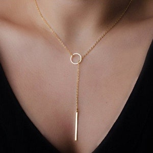 Peut inclure: Un collier doré avec une chaîne fine et un pendentif circulaire. Un pendentif en forme de barre verticale est suspendu au cercle, créant un design minimaliste et moderne. Le collier est présenté sur un fond sombre.
