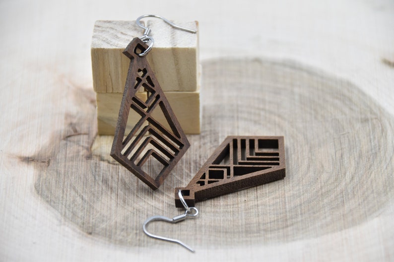 May include: Geometric wooden earrings with a dark brown finish. The earrings feature a unique open design with angular shapes and silver-colored ear wires. One earring is displayed separately, and the other is attached to a silver hook.