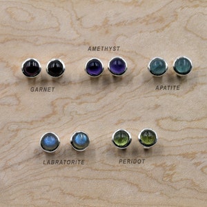 Peut inclure: Une collection de boucles d'oreilles à clous en pierres précieuses, dont grenat, améthyste, apatite, labradorite et péridot. Chaque paire présente une pierre ronde polie sertie dans une lunette en argent. Les noms des pierres sont étiquetés.
