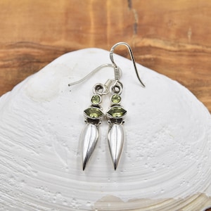 Peut inclure: Une paire de boucles d'oreilles en argent avec un design en forme de goutte. Chaque boucle d'oreille présente un élément argenté en forme de goutte d'eau, orné de deux pierres précieuses vertes. Les boucles d'oreilles sont présentées sur une surface blanche texturée.