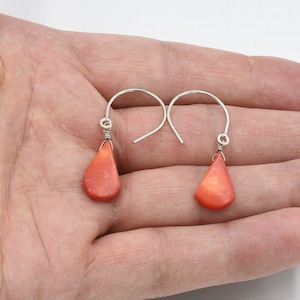 Peut inclure: Une paire de boucles d'oreilles en forme de goutte d'eau avec des pendentifs en pierre polie orange-rouge. Les boucles d'oreilles ont des crochets d'oreille argentés et sont attachées avec du fil d'argent.