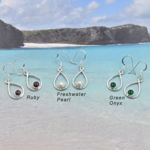 Peut inclure: Boucles d'oreilles argentées en forme de goutte d'eau avec des pierres de rubis, de perles d'eau douce et d'onyx vert. Chaque boucle d'oreille a une petite pierre ronde en bas. Sur fond de plage.