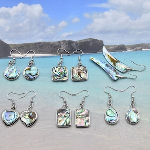 May include: A selection of abalone shell earrings in diverse shapes, such as round, square, heart, and teardrop. Each earring has a silver-tone metal frame and a unique iridescent pattern of blues, greens, and purples. The earrings are displayed against a beach backdrop.