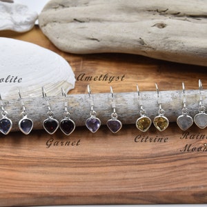Peut inclure: Une collection de boucles d'oreilles en forme de cœur avec des pierres précieuses, dont Lolite, Grenat, Améthyste, Citrine et Pierre de Lune Arc-en-ciel. Chaque boucle d'oreille a un crochet en argent et une pierre polie en forme de cœur. Les boucles d'oreilles sont exposées sur une surface en bois.