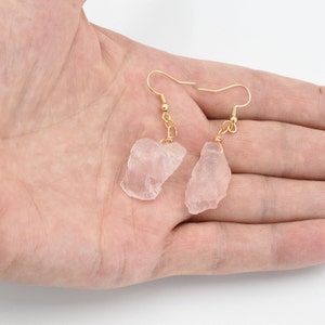 Peut inclure: Une paire de boucles d'oreilles en quartz rose. Chaque boucle d'oreille présente une pierre rose pâle taillée grossièrement, suspendue à un crochet doré. Les boucles d'oreilles sont présentées sur un fond neutre.