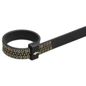 Könnte beinhalten: Schwarzer Ringgrößenmesser mit goldenen Zahlen und Markierungen. Der Ringgrößenmesser hat ein Band mit Zahlen von 44 bis 76 und einen Messstreifen. Ein Pfeil zeigt das Maß an.