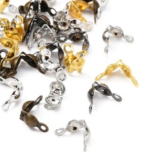 Könnte beinhalten: Eine Sammlung von Metall-Quetsch-Endstücken in verschiedenen Farben, darunter Gold, Silber und Bronze. Diese kleinen, gebogenen Teile haben an einem Ende eine Schlaufe und werden in der Schmuckherstellung verwendet, um die Enden von Kordeln oder Ketten zu sichern.