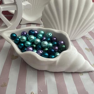 Könnte beinhalten: Eine weiße, muschelförmige Schale gefüllt mit türkisfarbenen, blauen und violetten Glasperlen. Die Schale steht auf einer rosa-weißen gestreiften Oberfläche.