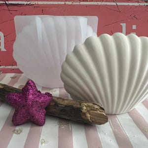 Könnte beinhalten: Ein weißer muschelförmiger Keramikbehälter und eine transparente muschelförmige Form. Ein pinkfarbener Glitzer-Seestern und ein Stück Treibholz befinden sich vor den Behältern. Der Hintergrund ist eine rosa-weiß gestreifte Oberfläche.