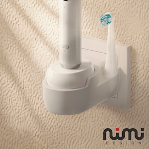 Peut inclure: Une brosse à dents électrique blanche et sa base de charge montées sur une prise électrique blanche. La brosse à dents a une tête de brosse bleue et blanche. La base porte l'inscription "Oral-B". Le texte "nimi DESIGN" est imprimé sous la prise.