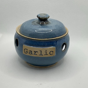 May include: A blue ceramic garlic keeper with a lid and a small knob. The keeper has a round shape with three circular holes and a label that reads "Garlic". The rim and the label have a tan colour.