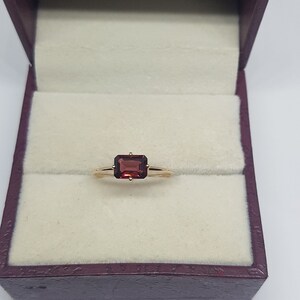 Könnte beinhalten: Ein goldener Ring mit einem roten rechteckigen Edelstein in einer Ringbox.