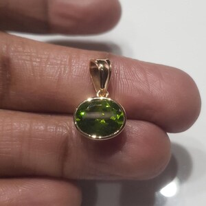May include: A gold pendant featuring a green oval gemstone. The pendant is resting on a finger.