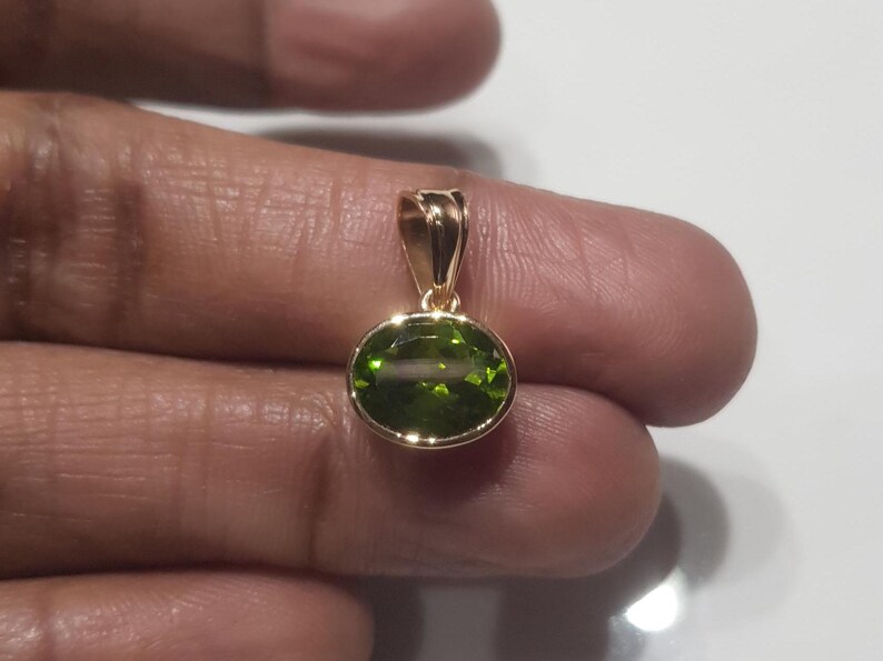 May include: A gold pendant featuring a green oval gemstone. The pendant is resting on a finger.