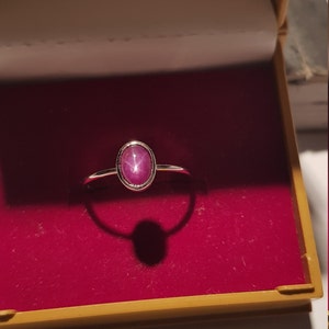 Puede incluir: Un anillo de plata con una piedra preciosa rosa ovalada en forma de estrella. El anillo está engastado en un diseño simple y elegante y se muestra en una caja de joyería forrada de terciopelo rojo con un exterior dorado. La gema tiene un efecto de estrella.