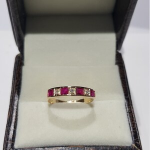 May include: A gold band ring with a row of red square-shaped gemstones and smaller clear gemstones.