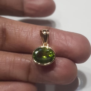 May include: A gold pendant featuring a green oval gemstone. The pendant is held on a finger.