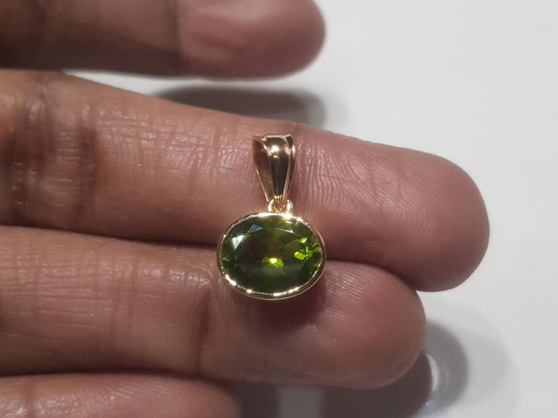 May include: A gold pendant featuring a green oval gemstone. The pendant is held on a finger.