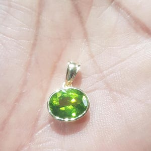 May include: A gold pendant featuring a green oval gemstone. The pendant is resting on a hand.