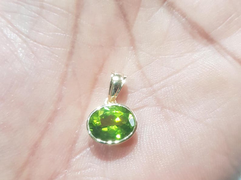 May include: A gold pendant featuring a green oval gemstone. The pendant is resting on a hand.