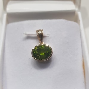 May include: A gold pendant featuring a green oval gemstone. The pendant is displayed in a white box.