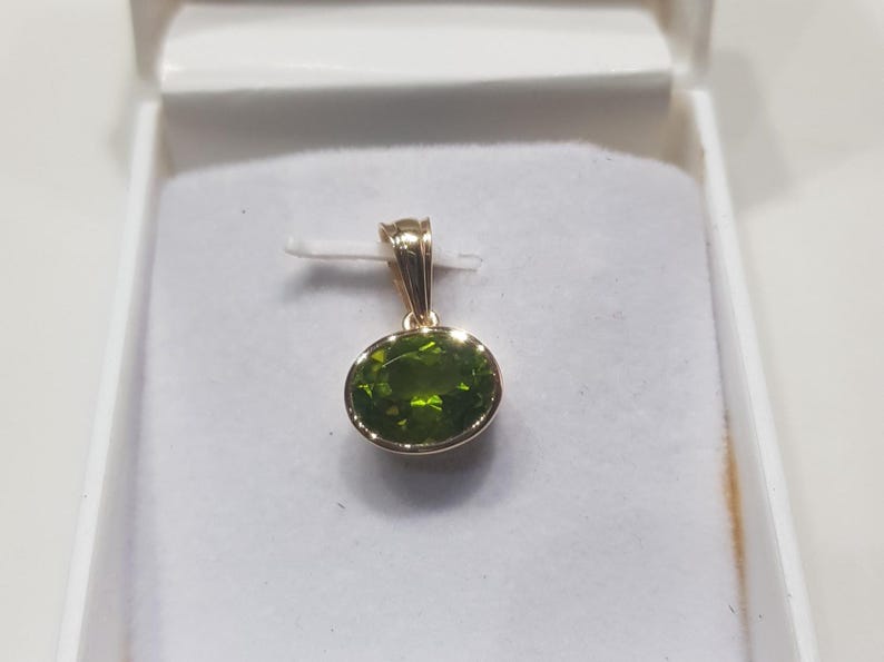 May include: A gold pendant featuring a green oval gemstone. The pendant is displayed in a white box.