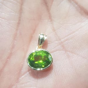 May include: A gold pendant featuring a green oval gemstone. The pendant is resting on a hand.