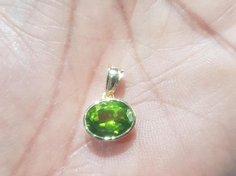 May include: A gold pendant featuring a green oval gemstone. The pendant is resting on a hand.