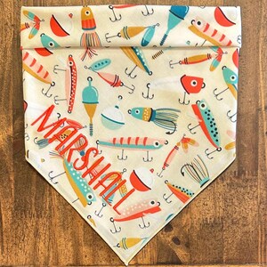 Puede incluir: Un pa&ntilde;uelo triangular para mascotas con tela de color crema y un patr&oacute;n repetitivo de se&ntilde;uelos de pesca, anzuelos y boyas de colores. El nombre MARSHALL est&aacute; bordado con hilo rojo anaranjado intenso en diagonal sobre el centro.