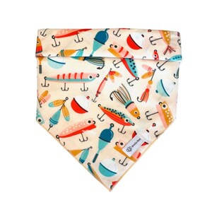 Puede incluir: Un pa&ntilde;uelo triangular para mascotas con fondo crema que presenta un patr&oacute;n colorido de se&ntilde;uelos de pesca, anzuelos y boyas en tonos rojos, azules, naranjas y amarillos. Una peque&ntilde;a etiqueta de tela blanca con el texto Stella Mae est&aacute; cosida.